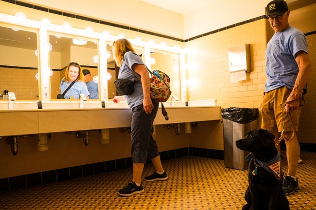 A woman turns on the automatic faucets in the bathroom while a man holds the leash for the black puppy. The puppy sits and watches her.