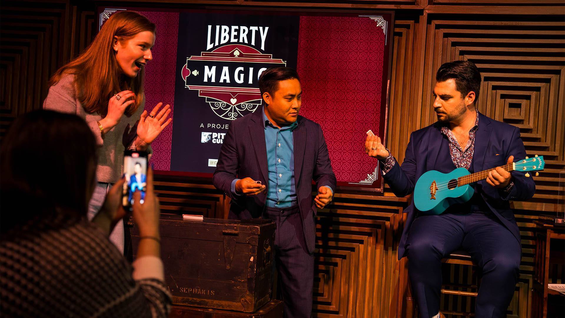 Three people interacting on a small stage. The person on the right is holding a small, teal guitar. The other two people look like they're guessing something.