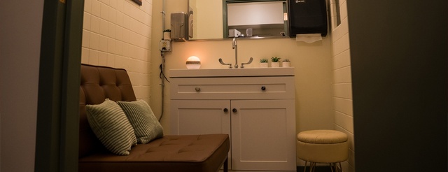 A close up of white vanity and sink, a mirror, and the chocolate brown lounge chair with green pillows.