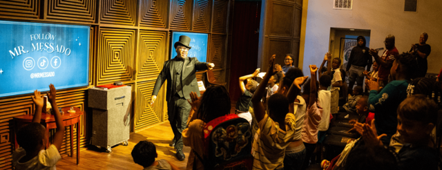 Joshua Messado in his magician's uniform: black top hat, black vest, and a black suit, smiles while pointing at a group of kids with their hands raised.