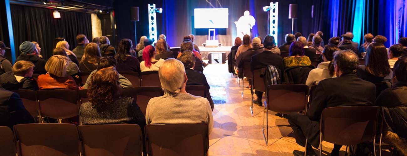 a crowd of seated people all watch a speaker on a stage in front of them