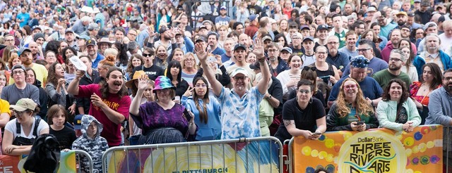 A large, diverse crowd stands outdoors behind barricades at the Three Rivers Arts Festival in Pittsburgh, smiling and waving at the camera on a bright day, with festival banners visible in front.