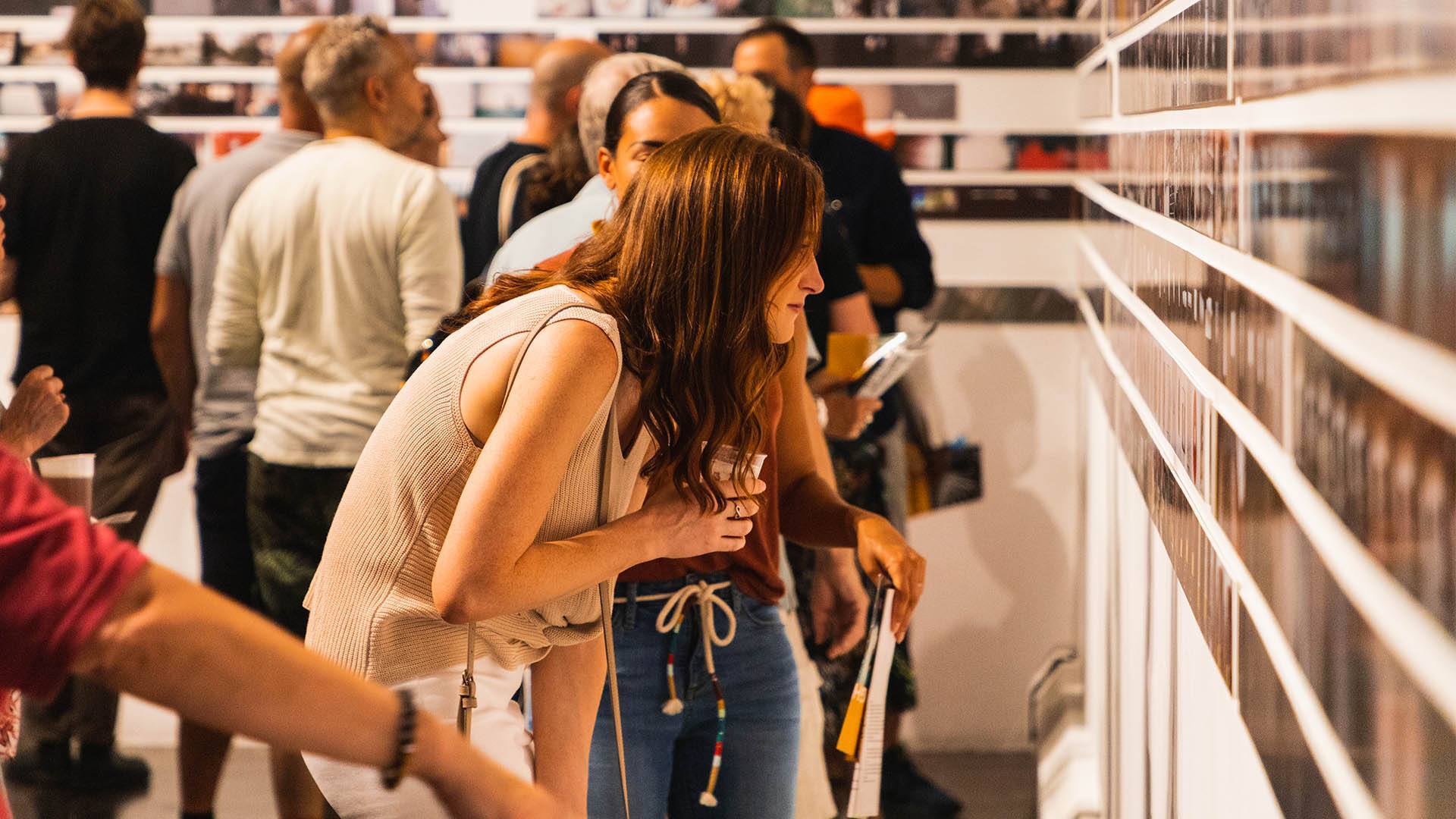 A woman with shoulder length, brown hair holding a drink. She is bent over slightly looking closely at artwork. Many other people converse in the background.
