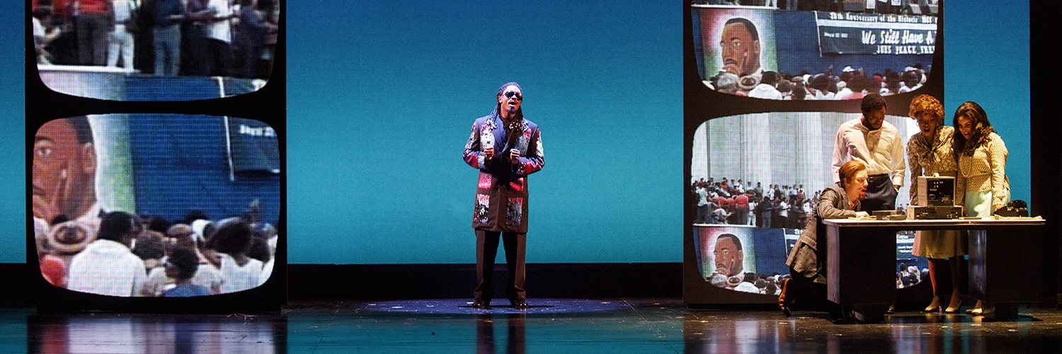 Actor playing Stevie Wonder stands center stage. Martin Luther King, Jr. projections are featured on either side.