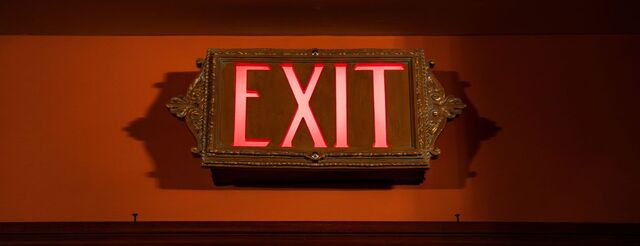 A close up of an exit sign with glowing red letters in an ornate, brass colored casing.