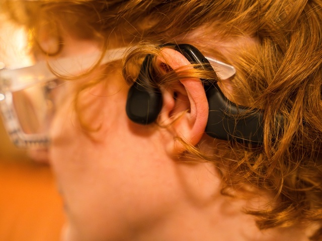 A woman sits in a theater seat wearing glasses, a hearing aid, and bone conduction headphones.