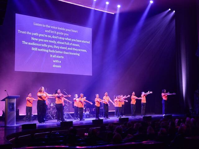 A group of middle school students in matching orange shirts sing on stage under purple lights.
