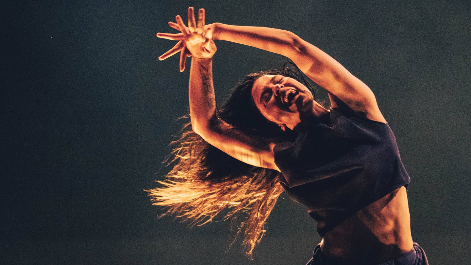 A muscular dancer with long, flowing hair. They have their hands extended above their head with fingers spread and leaning their body to their right. They are smiling but look like they're slightly in pain.