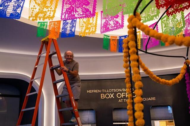 A man stands on a ladder with multi-colored papel picado above his head.