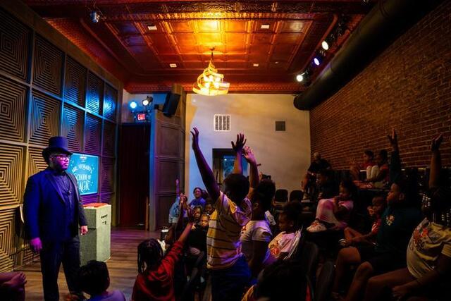 Messado looks out on the crowd as children raise their hands, some are standing up.