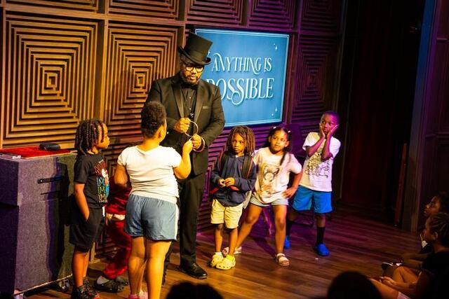Messado holds up a ring while a child holds up the other ring that it is linked to. A few other children stand on stage watching the trick unfold.