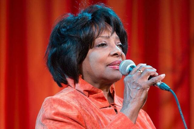 A women in a red jacket and a red background holds up a microphone.