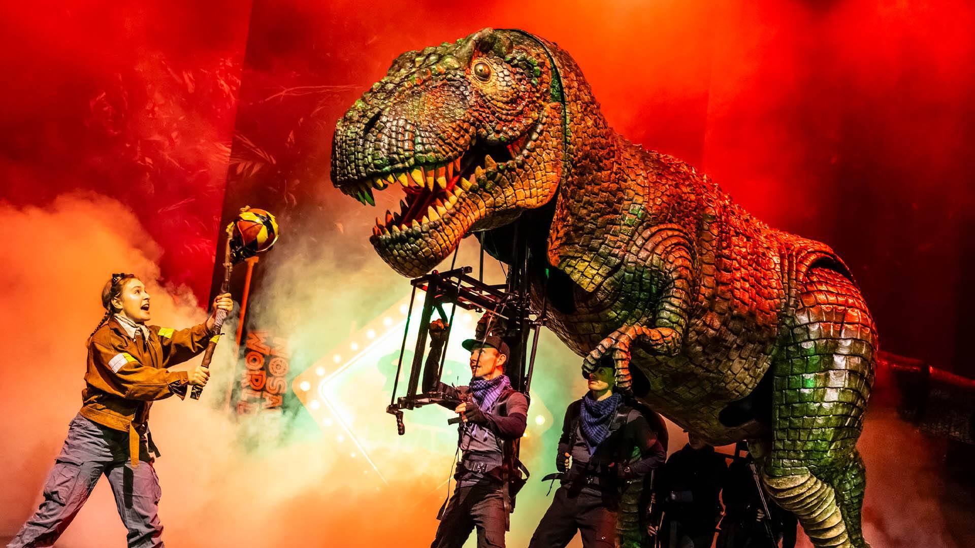 A large, realistic T-Rex puppet is operated by several performers on stage, with dramatic red lighting and smoke effects. A person in costume faces the dinosaur, holding a prop, creating an intense, theatrical scene.