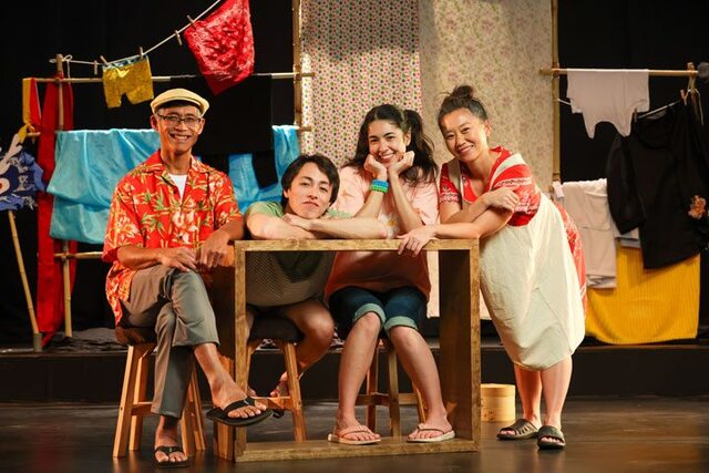 An Asian family - two adults, one boy, and one girl, sit at a table and look at the camera. Behind them are clothes hung up on a clothes line.