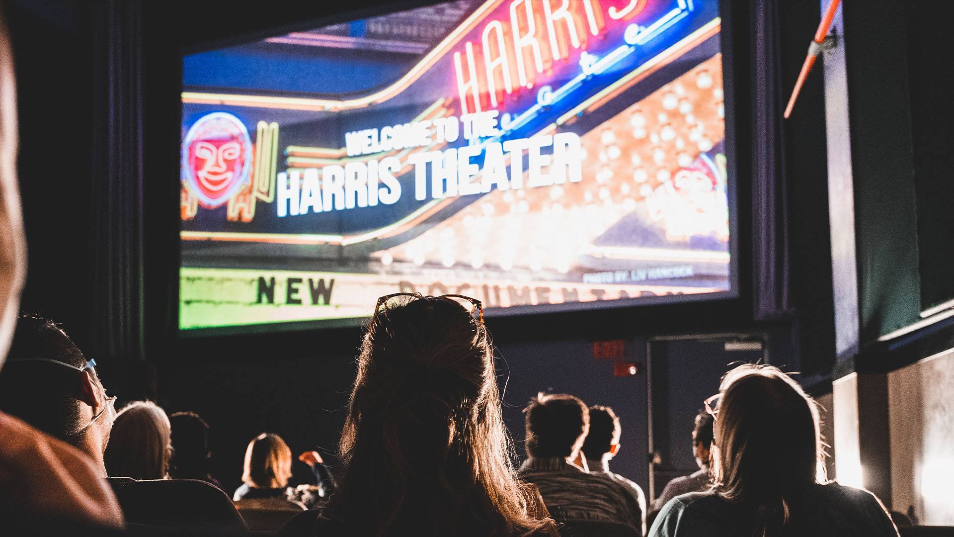People sitting in a movie theater watching a film. The screen says 'Welcome to the Harris Theater'.