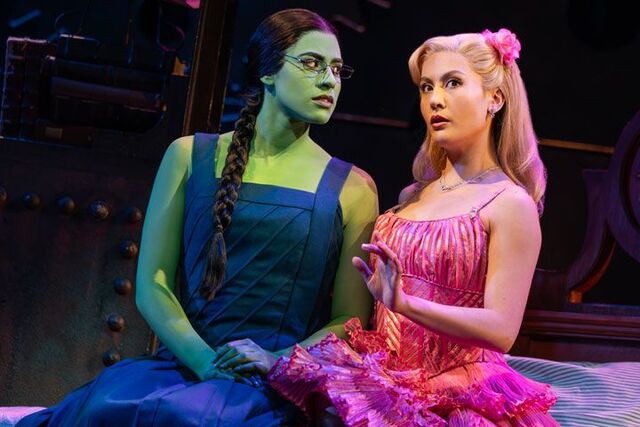 Glinda in a pink ball gown and Elphaba and in black dress and witches sit with one another.
