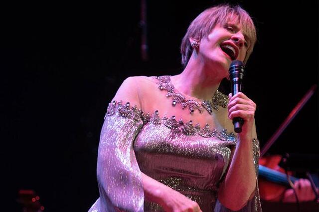 Patti Lupone, wearing a silver sparkling dress, sings into the microphone.