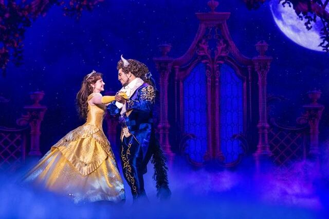 A woman in yellow ball gown and a beast in a blue suit with intricate detailing dance hand in hand. Behind them in a set background with a moon and an ornate gate. At their feet is a layer of fog.