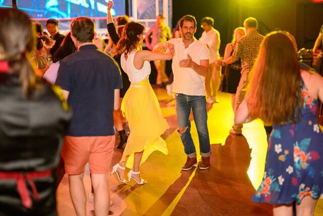 Several partners dance together. In focus, a man in jeans gets ready to turn a woman in a yellow skirt.