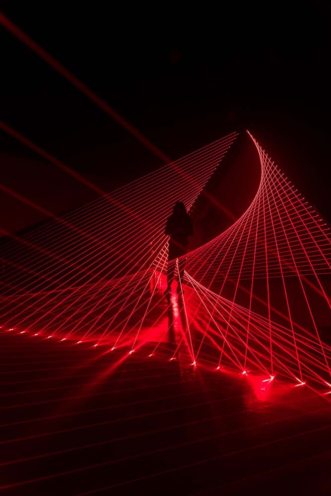  A detail view of Double Gemini. A silhouette of a person stands among red lasers that crisscross in front of and behind them