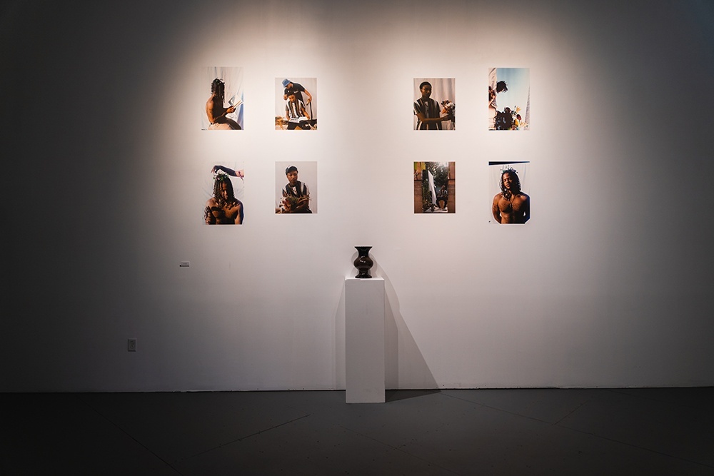 eight photograph prints hand on a white wall. a brown vase stands on a plinth in front of them