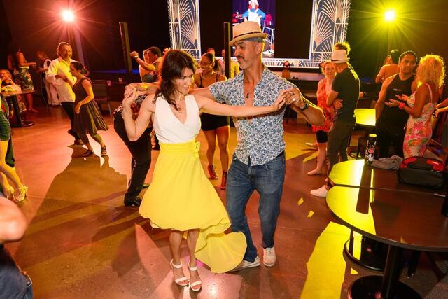 A man and woman dance together in a lively, colorful room. The woman wears a white top and yellow skirt, while the man wears a hat and patterned shirt. Other people dance and watch in the background under bright lights.