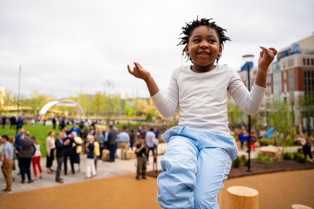 A smiling child in a white shirt and blue pants jumps or poses outdoors, with a park and a crowd of people in the background on a cloudy day.