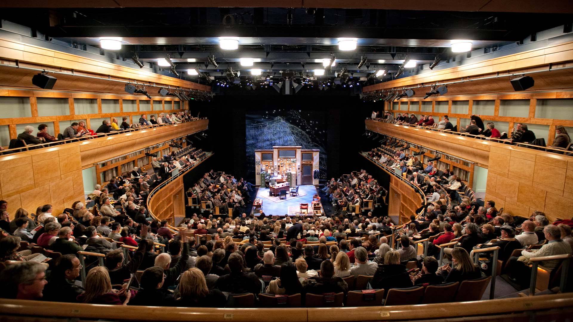 A large audience fills a modern, wooden theater with two balconies, watching a stage set for a play. The stage features furniture and a backdrop resembling a room.