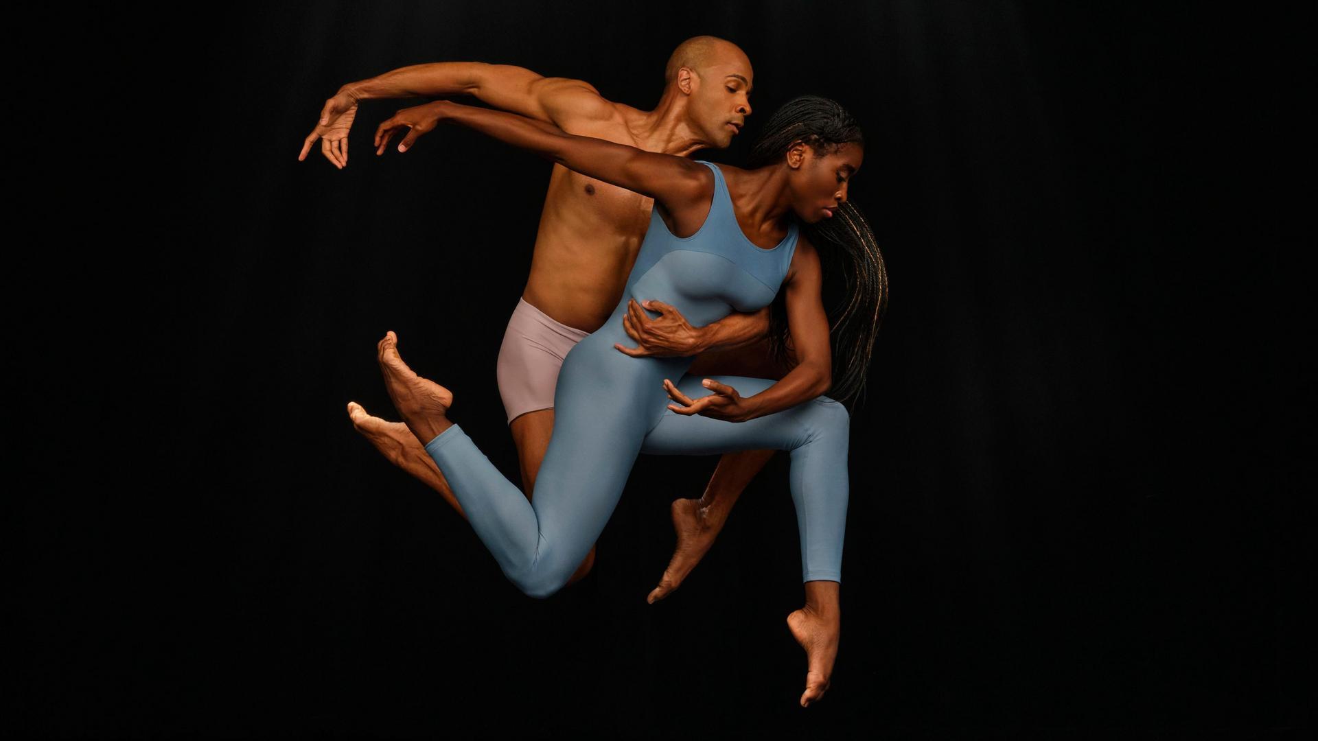 Two dancers perform a dynamic leap together against a black background. The woman in a blue bodysuit is in front, with the man behind her, supporting her mid-air. Both have focused expressions and extended limbs.