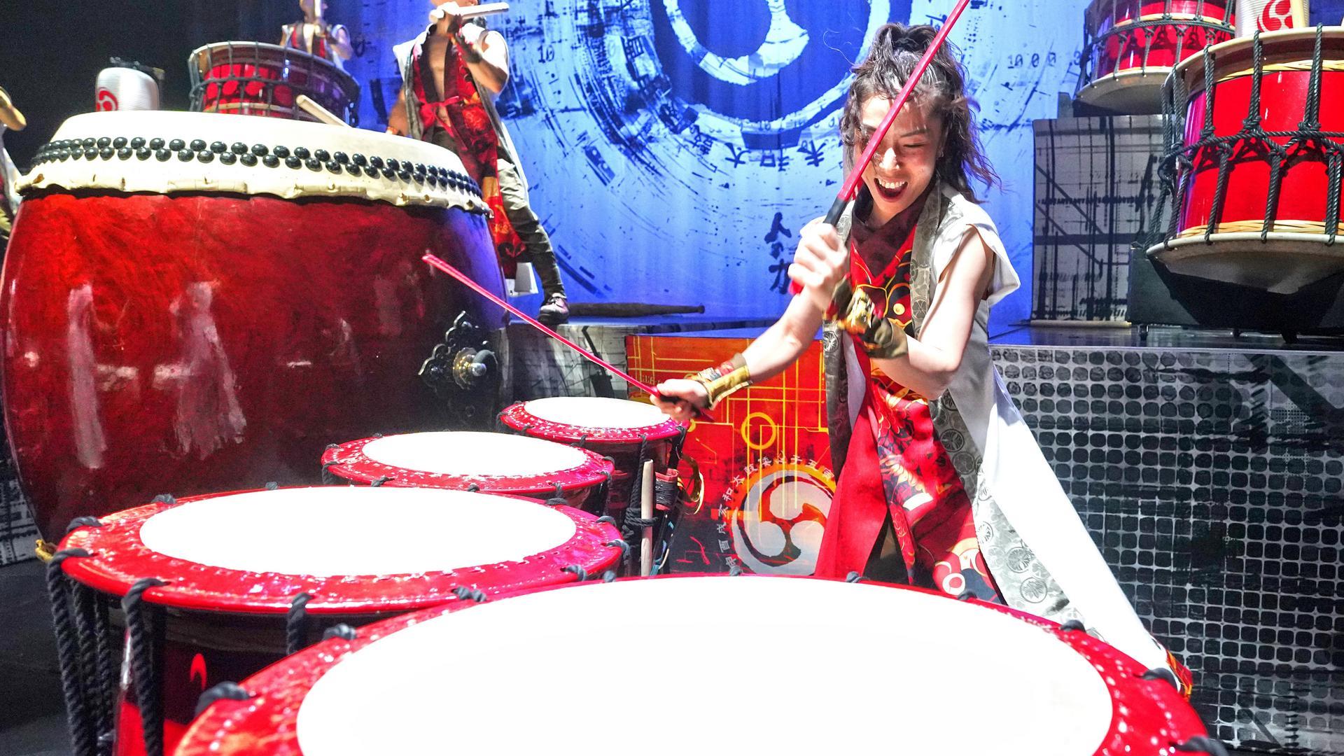 A joyful woman in a colorful costume plays multiple traditional Japanese taiko drums on stage, surrounded by large drums and a blue patterned backdrop.