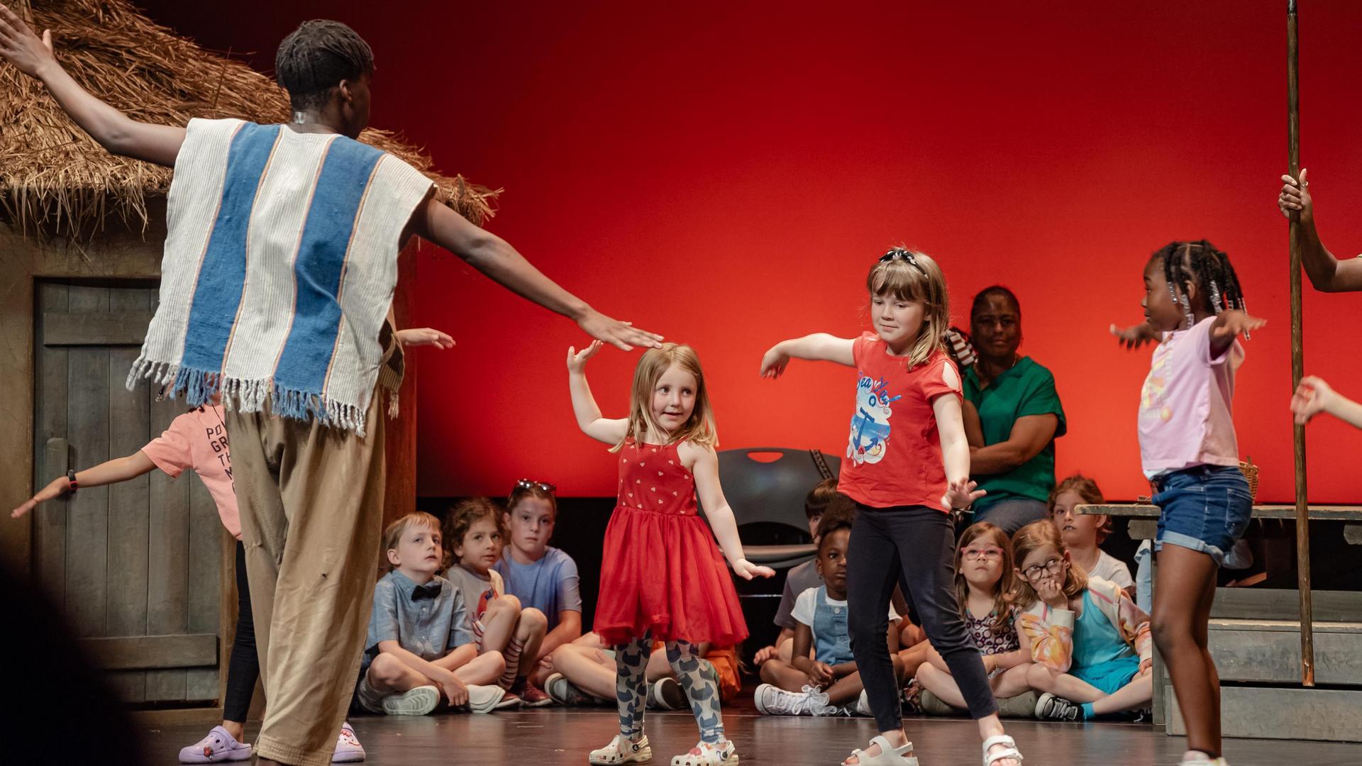 A group of young children stand on stage learning a dance from an adult instructor in a striped vest, with other kids seated behind them and a red backdrop.