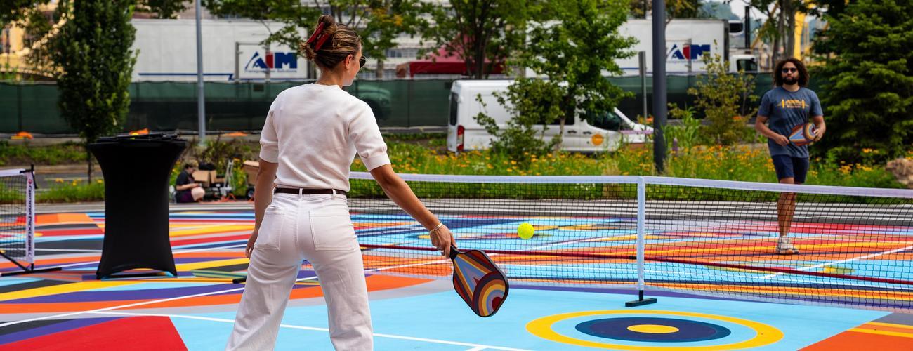 A woman in white plays pickleball on a colorful outdoor court with a man. Trees, flowers, and parked vehicles are in the background.