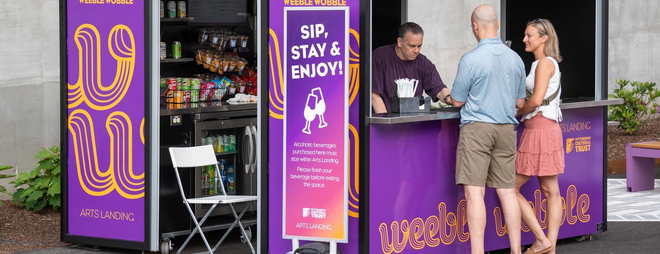 A man serves a couple at a purple outdoor drink kiosk with snacks on shelves. Signs say Sip, Stay & Enjoy! and promote keeping drinks at the venue.