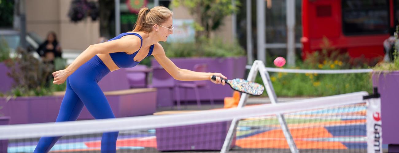 A woman in a blue athletic outfit plays pickleball outdoors, reaching with her paddle to hit a ball over the net on a colorful court.