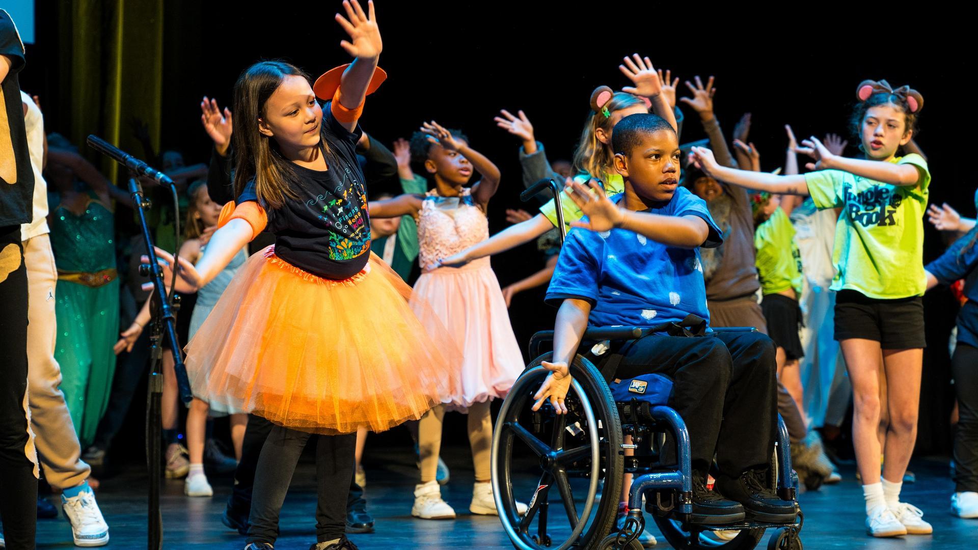 Children, including a boy in a wheelchair, perform an energetic group dance on stage. They wear colorful costumes, and everyone has their arms raised enthusiastically under bright stage lights.