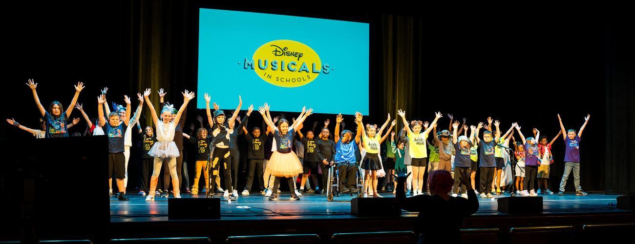 A large group of children on a stage with their arms raised in front of a blue screen that reads Disney Musicals in Schools.