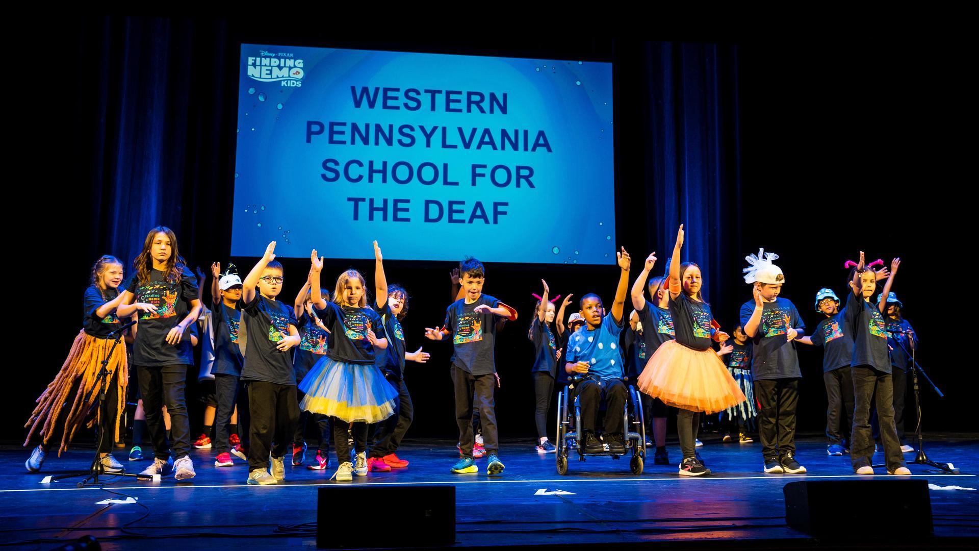 A group of children in costumes perform on stage under a screen that reads Western Pennsylvania School for the Deaf. Some raise their hands, and one child uses a wheelchair. Blue stage lighting highlights the scene.
