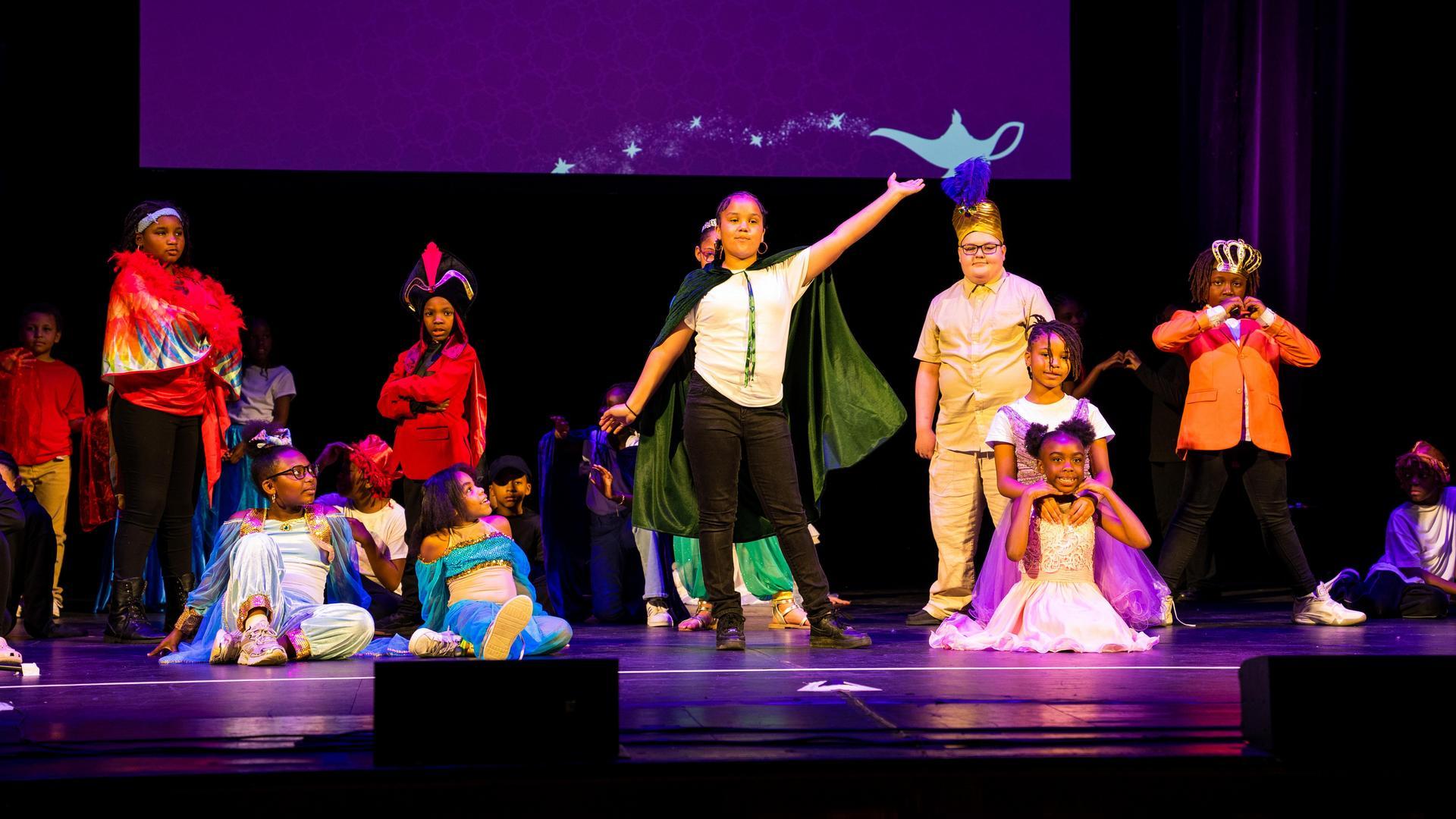 Children in colorful costumes perform on stage, some standing, some sitting, under purple lighting. A projection of a magic lamp is visible on the screen behind them. The scene suggests a theatrical or school play.