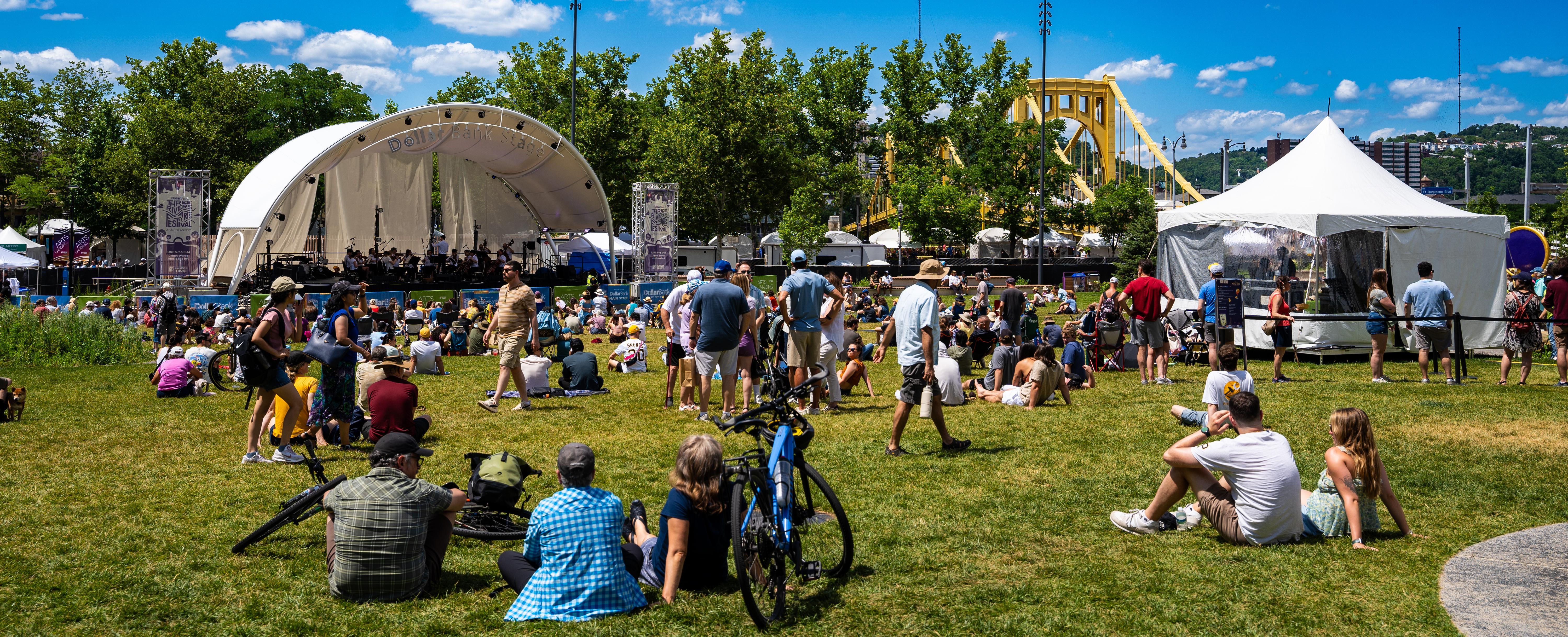 People sitting on the grass at an outdoor park event with a white performance stage, a white canopy tent, and a yellow bridge in the background.